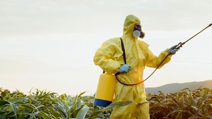 Trabajador agrícola con traje protector y mascarilla rociando pesticidas en un campo de cultivo