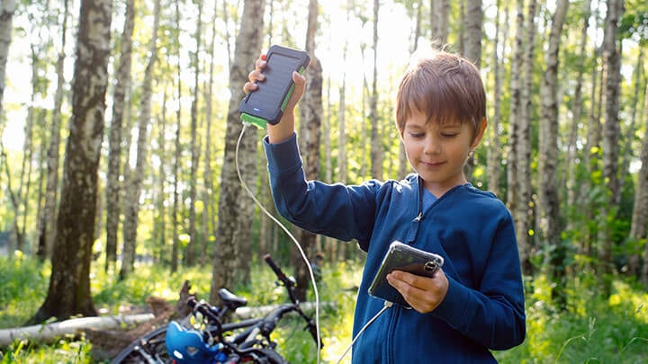 Un joven con una chaqueta azul con cremallera en un sendero para bicicletas con una bicicleta al fondo sosteniendo un pequeño dispositivo de panel solar conectado a un teléfono